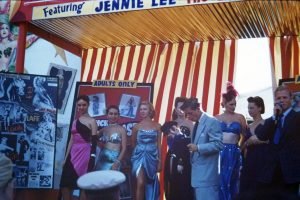 Photo of bally for Jennie Lee's show at the Canadian National Exhibition, 1956. Image shows several female performers lined up next to a collage of images of Jennie Lee. A red striped tent wall is visible behind them. To the right is the show's talker, a man in a blue suit with a microphone. In the foreground, a man in a military cap looks on.