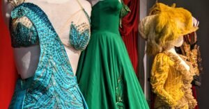Detail of Built to Bare showing three costumes, a beaded top depicting a peacock, a green dress, and a yellow dress with feathered hat.