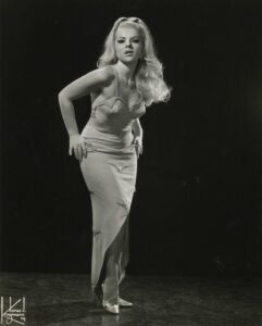 Black and white promotional photo of Val Valentine. She stands facing the vcamera against a black background, hands on hips, torso pitched forward slightly. She wears a light-colored dress cut low in front and slit to her knee. Her hair is blonde and flows over her left shoulder from a high ponytail.