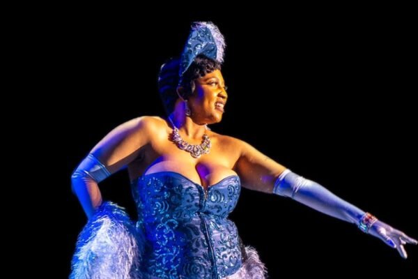 A performer in a blue corset and gloves extends an arm out while smiling.