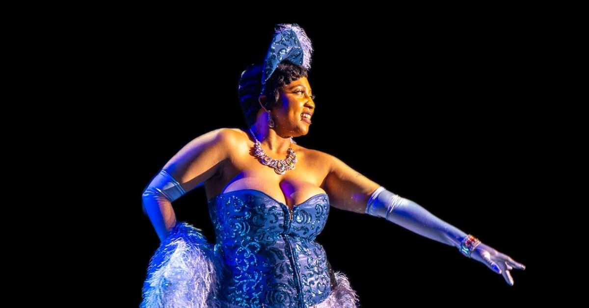 A performer in a blue corset and gloves extends an arm out while smiling.