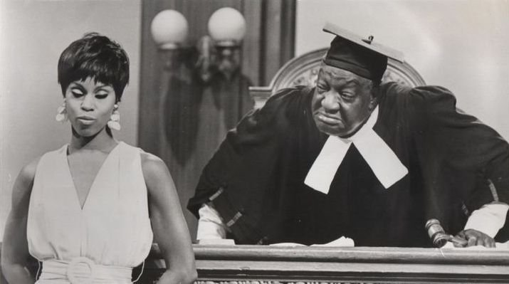 Dressed as a judge, Dewey "Pigmeat" Markham scowls over the bench at a woman.