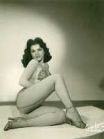 Black and white promotional photo of Val Valentine. She is reclining on a pedestal, right leg tucked to the side and left leg bent in front of her, covering much of her torso, which is turned to her left. She looks over her right shoulder directly at the viewer. Her dark hair is cut at shoulder length and framed her face in large waves at both sides. She wears fishnet stockings and high heels with a sparkly bra top.