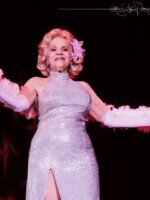 Color photo of Val Valentine performing at the 2018 Burlesque Hall of Fame Weekender. She wears a form-fitting white dress covered with closely-spaced silver sequins, elbow-length white gloves, a white boa draped over her arms and behind her back, and a pink flower in her hair. The image is from a low angle; she is facing towards the camera but looking over it. Her blonde hair is cut to about her chin and arranged in large curls. Color photo of Val Valentine performing at the 2018 Burlesque Hall of Fame Weekender. She wears a form-fitting white dress covered with closely-spaced silver sequins, elbow-length white gloves, a white boa draped over her arms and behind her back, and a pink flower in her hair. The image is from a low angle; she is facing towards the camera but looking over it. Her blonde hair is cut to about her chin and arranged in large curls.