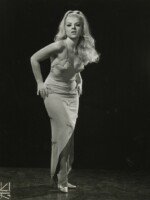 Black and white promotional photo of Val Valentine. She stands facing the vcamera against a black background, hands on hips, torso pitched forward slightly. She wears a light-colored dress cut low in front and slit to her knee. Her hair is blonde and flows over her left shoulder from a high ponytail.